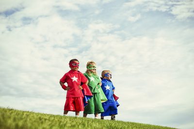 3 Kinder in Superheld*innenkostümen stehen auf einem Hügel und schauen kämpferisch in eine Richtung